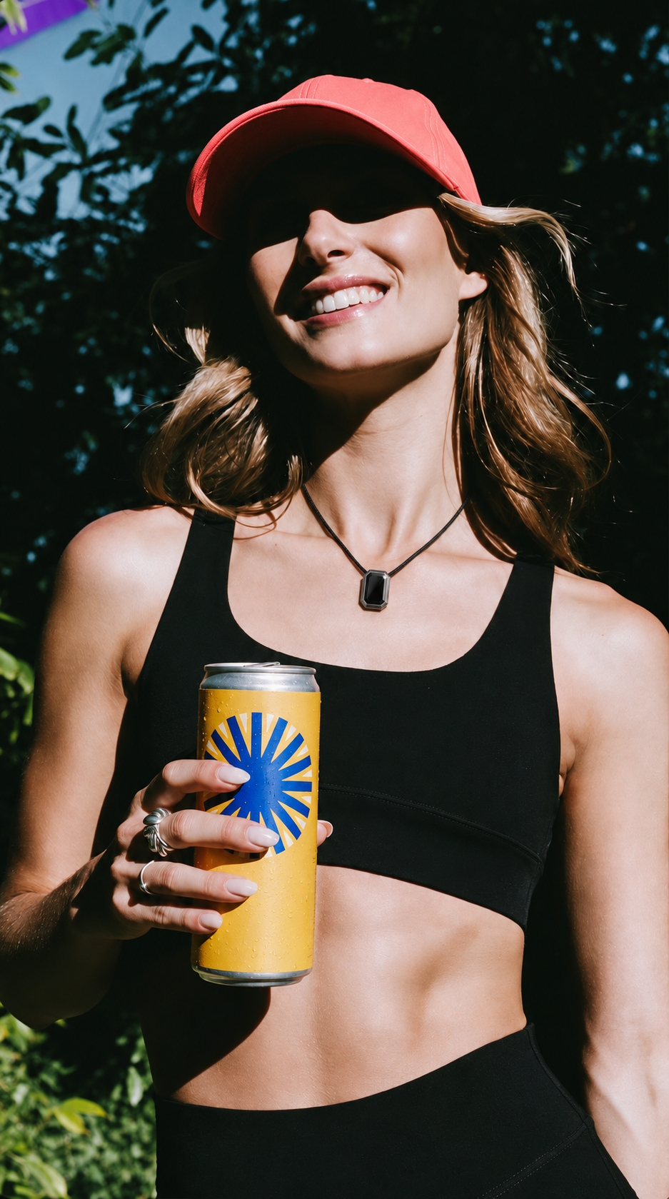A woman outdoors in a coral cap, wearing the ORIN pendant and holding a drink.